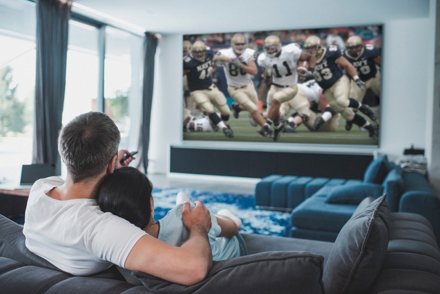 a couple lounges on the couch in front of a large screen TV playing a football game